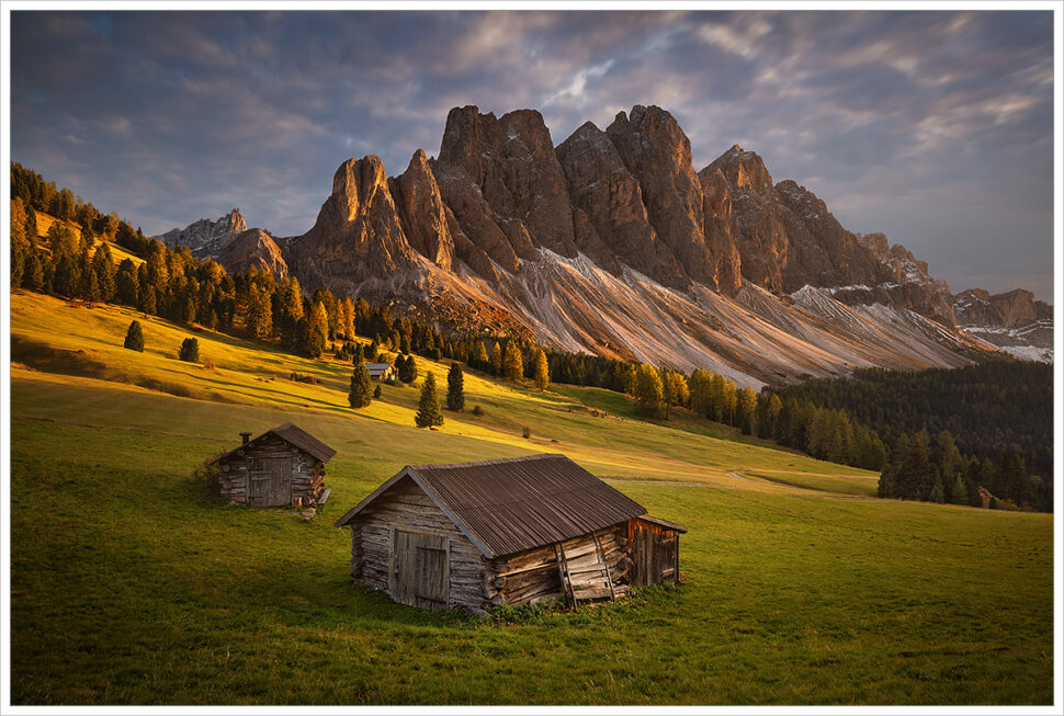 Podzimní večer v italských Dolomitech. V popředí jsou dva staré dřevěné seníky a v pozadí večerním sluncem nasvícené horské štíty Dolomit - Fotografický workshop Čarovné podzimní Dolomity