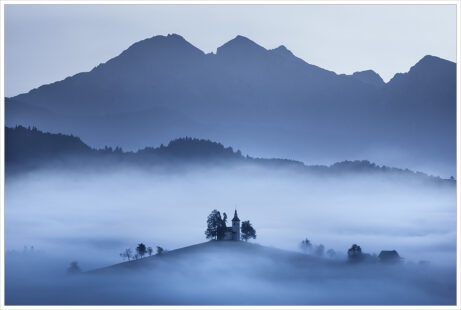Hornatá krajina Slovinska před východem slunce. Z&nbsp;ranní mlhy vystupují horské horizonty, klasický Slovinský kostel a&nbsp;několik stromů - Fotografický workshop Pohádkové podzimní Slovinsko